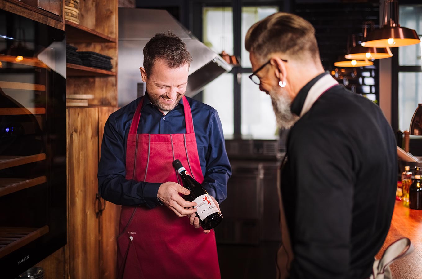 le tablier | zwei Männer in einer Küche mit einer Weinflasche und Schürzen