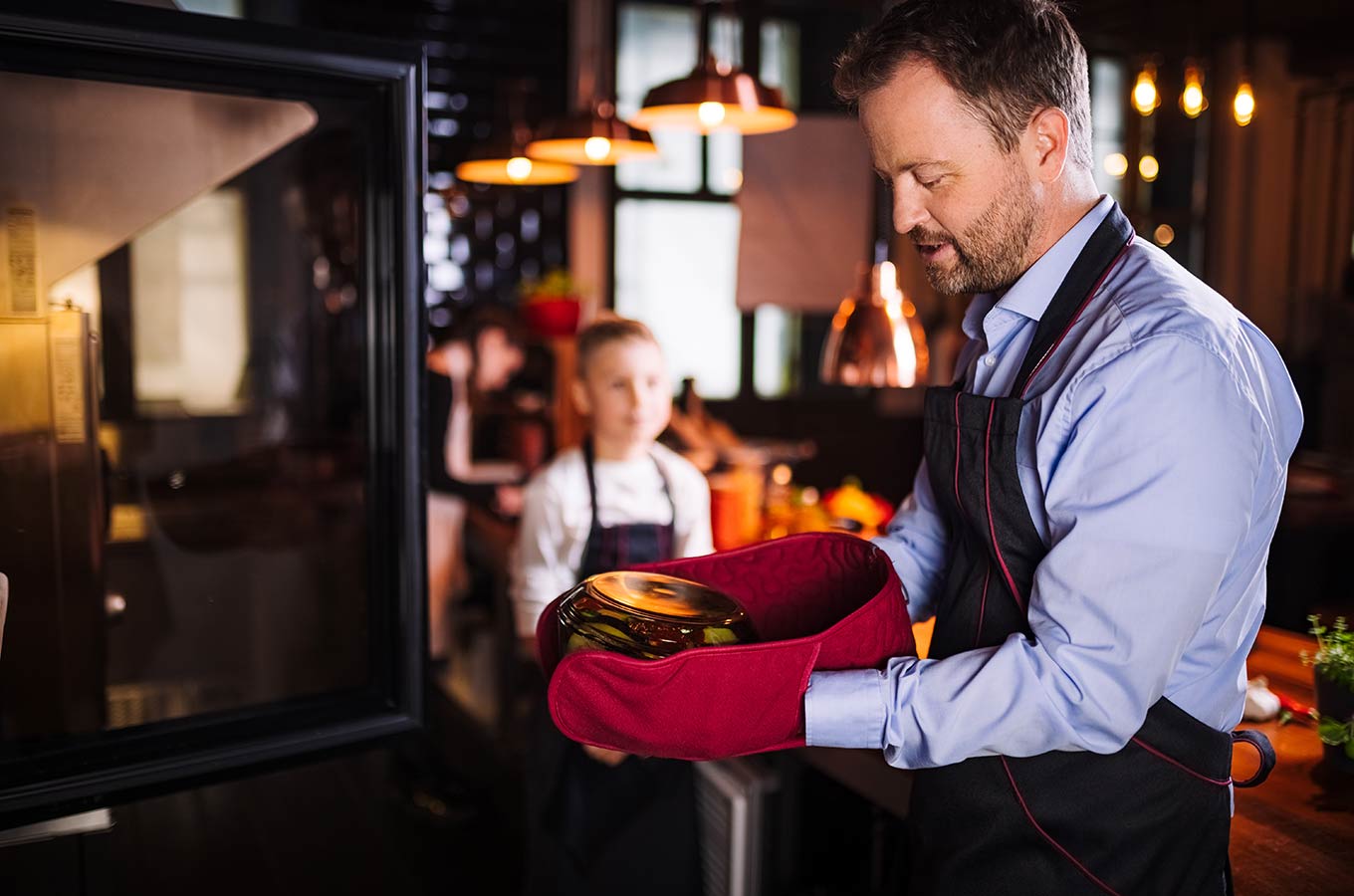 le tablier | Mann mit schwarzer Schürze und Topflappen vor einem Ofen stehend