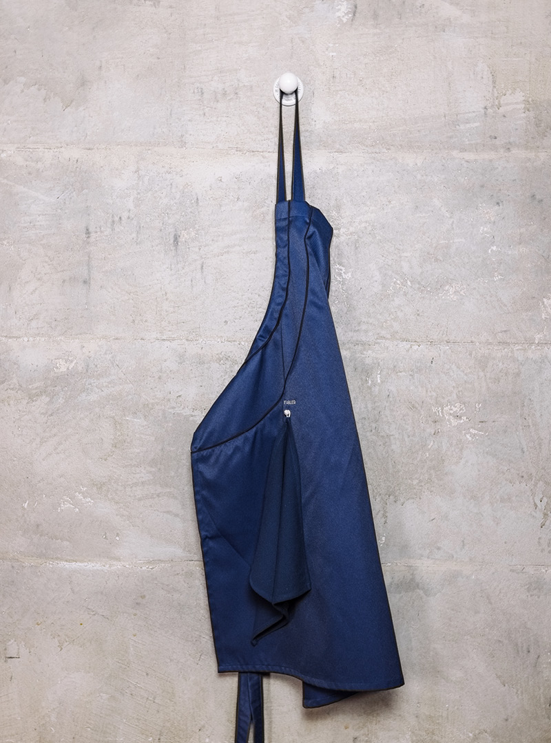 le tablier | dunkelblaue Schürze mit Abtrockentuch an weißem Haken an Betonwand
